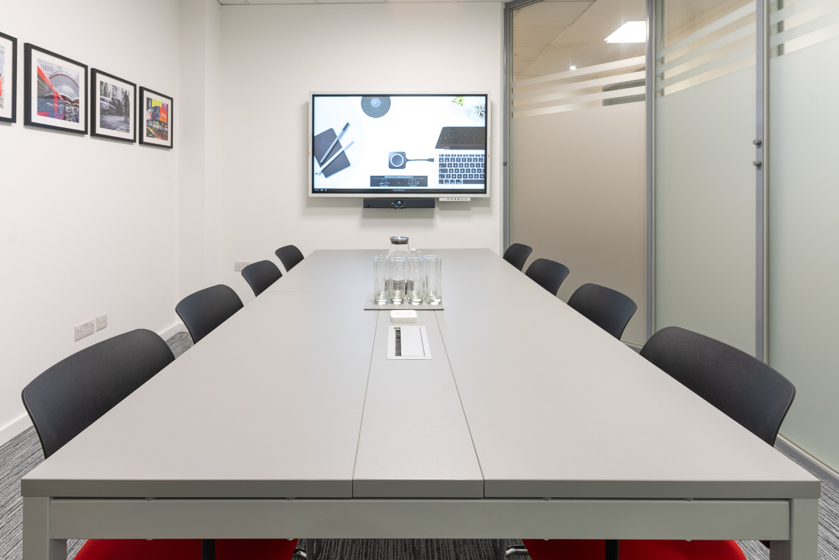 Meeting room with light grey walls and grey carpet floor tiles, black meeting table, 8 black and red meeting chairs, large display screen with video conferencing unit, and framed photographs hanging on wall.