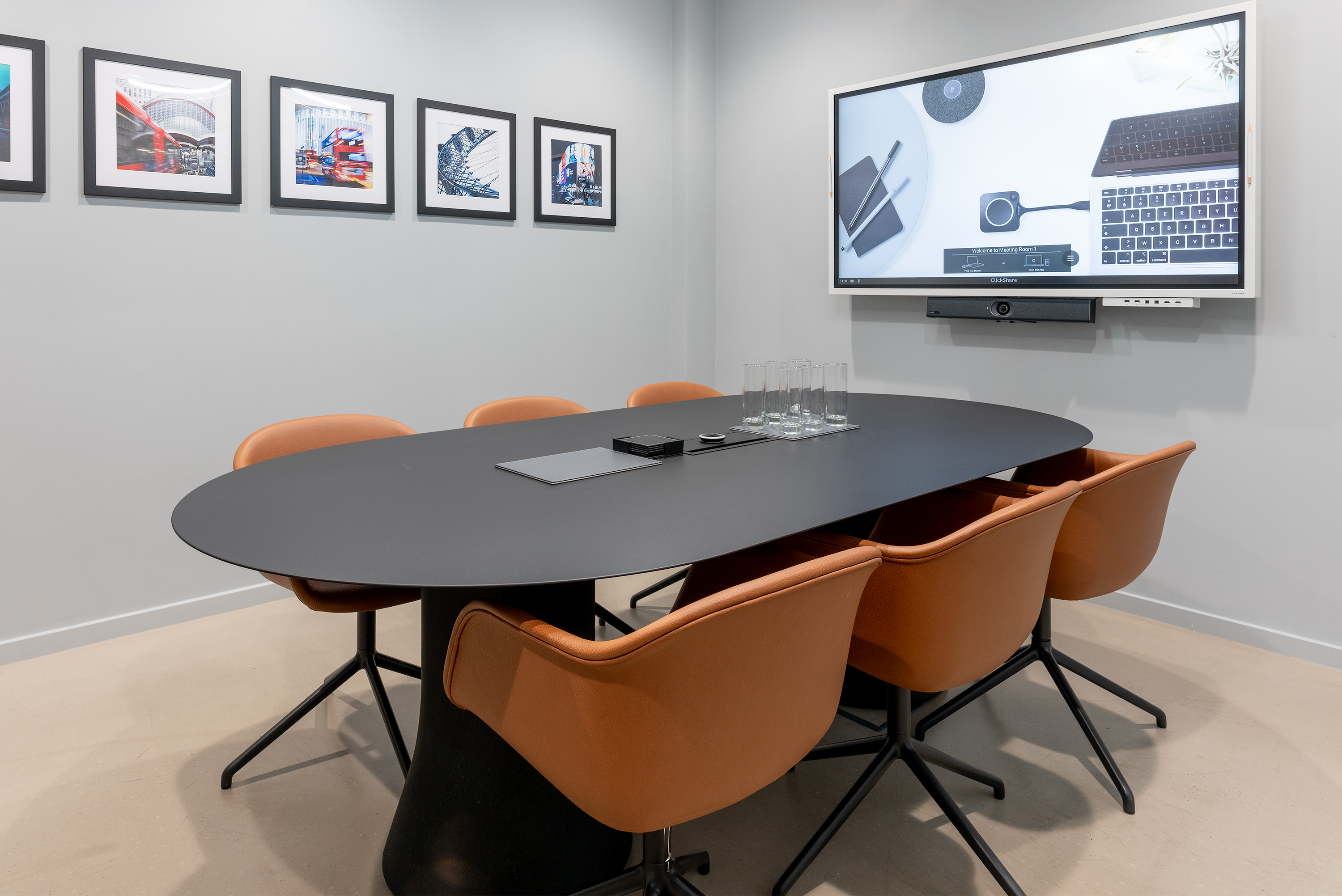 Meeting room with light grey walls and concrete floor, black meeting table, 6 brown tub chairs, large display screen and video conferencing unit, and framed photographs hanging on wall.