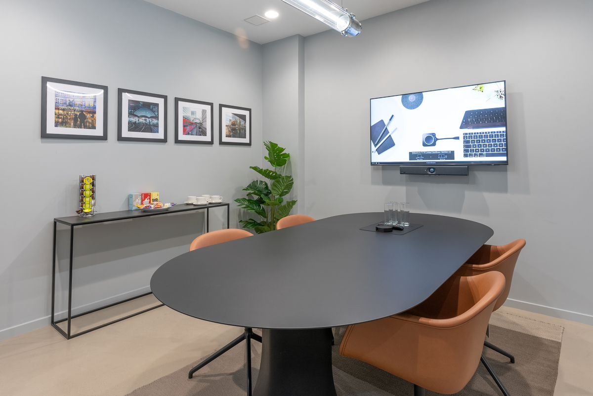 Meeting room with light grey walls and concrete floor, black meeting table, 4 brown tub chairs, large display screen and video conferencing unit, black credenza with tea and coffee accessories, and framed photographs hanging on wall.