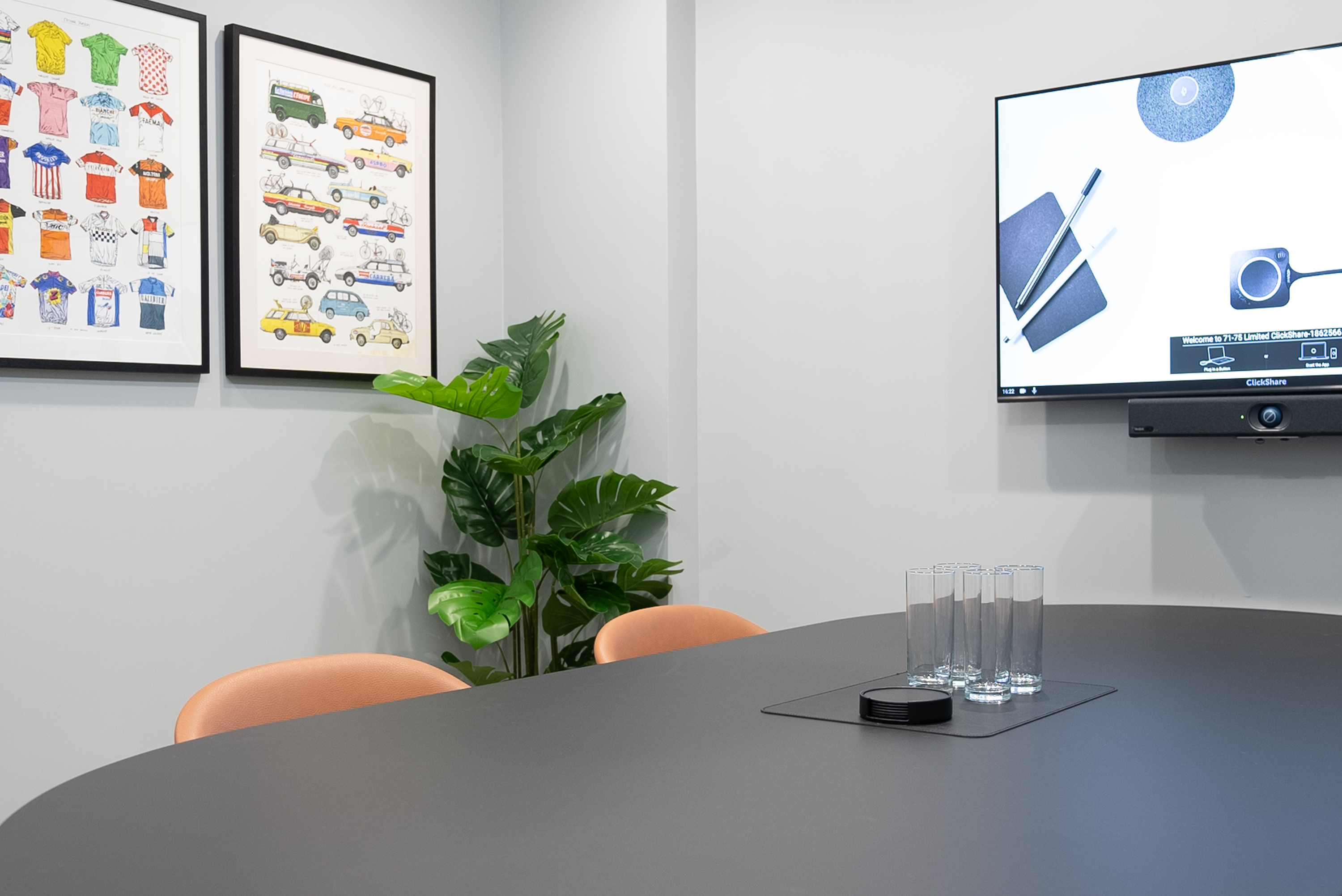 Meeting room with light grey walls and concrete floor, black meeting table, 4 brown tub chairs, large display screen with video conferencing unit, black credenza with tea and coffee accessories, and framed photographs hanging on wall.