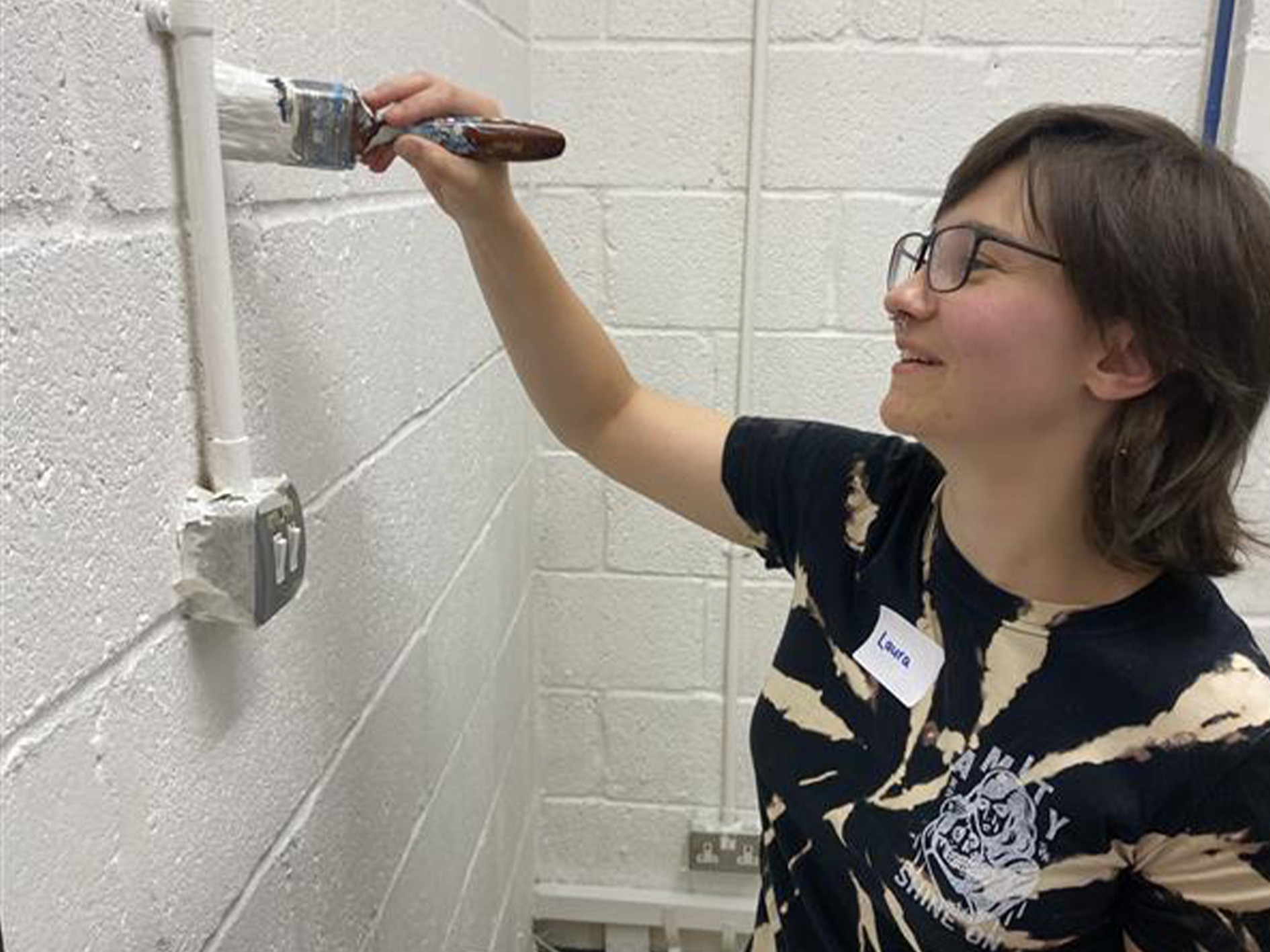 A woman wearing glasses, a black and beige tie-dye T-shirt, and a name tag reading 'Laura' is painting a white brick wall with a paintbrush. She is smiling while applying fresh white paint to a pipe. The electrical socket nearby is covered with tape to protect it from paint. The setting is a community project area.