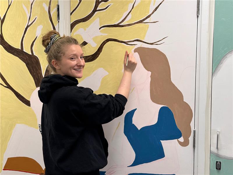 A person with blonde hair tied up in a bun, wearing a black hoodie, is painting a mural on a white wall. They are smiling at the camera while holding a paintbrush. The mural features a stylised figure of a woman in a blue dress with brown hair, as well as a large tree with dark brown branches on a yellow background. The setting appears to be an indoor space with a door integrated into the mural.