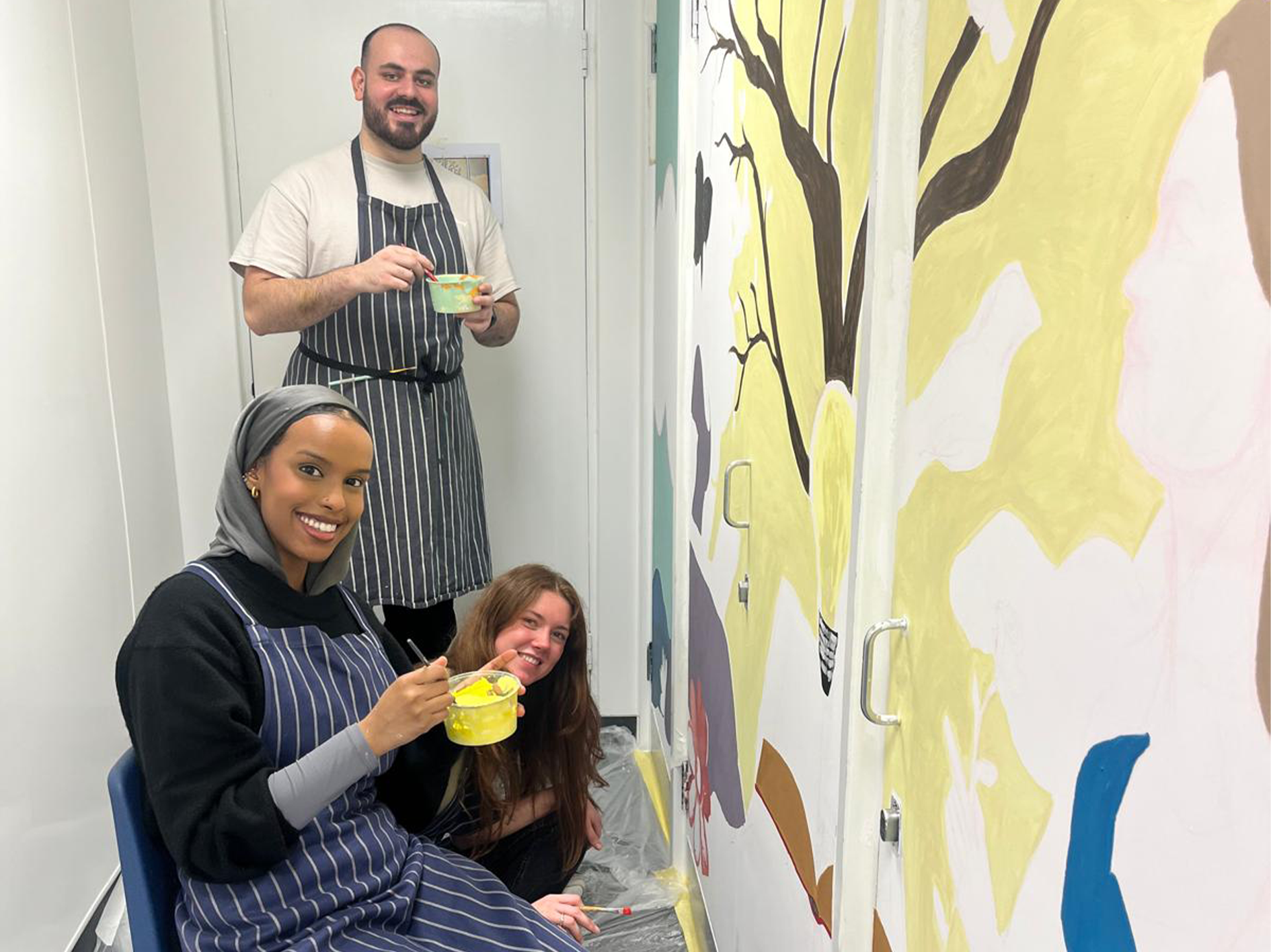 Three people painting a mural on a white wall in a narrow room. The mural features a large tree with dark brown branches and a yellow background resembling a light bulb. The individuals are wearing striped aprons and holding small containers of yellow paint with brushes. The person in the foreground, wearing a gray hijab and black clothing, is sitting on a chair and smiling at the camera. Another person with long brown hair is crouching beside her, also smiling. A third person, standing in the background, has a beard and is wearing a light-colored shirt under the apron while holding a paint container. The floor is covered with plastic sheeting.