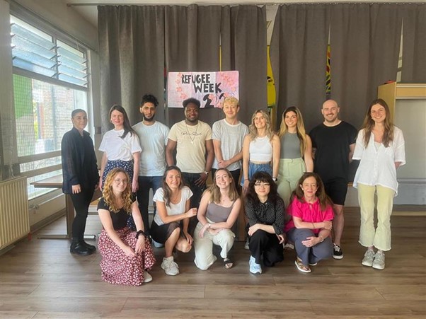 A group of thirteen people posing together in a well-lit room with wooden flooring. They are standing and kneeling in front of a hand-made sign that reads REFUGEE WEEK' with decorative elements. The group consists of individuals with diverse appearances, wearing a mix of casual and semi-formal outfits. They are smiling. Behind them, grey curtains cover parts of the wall, with some yellow and black decorations partially visible on the sides. The room has large windows with horizontal blinds on the left, allowing natural light to enter.