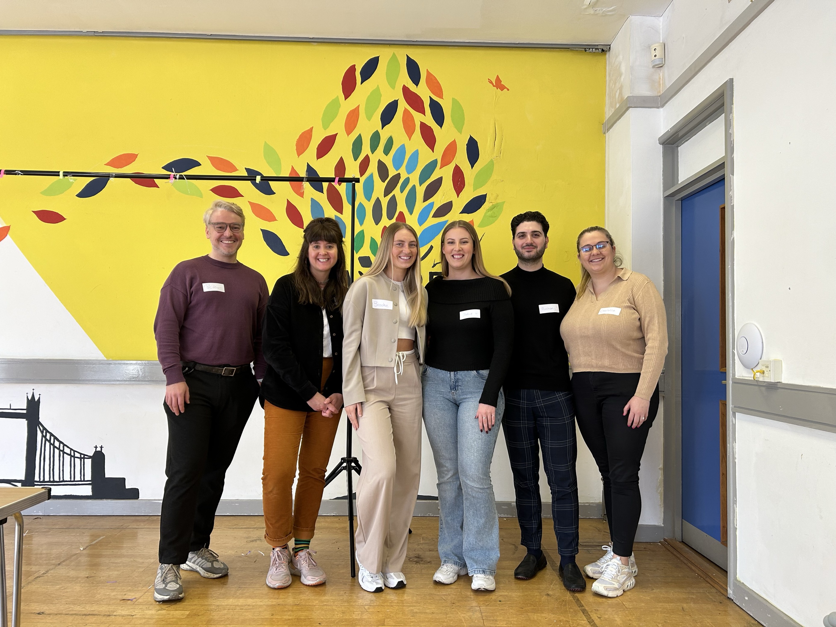 A group of six people stands together in a classroom at a corporate volunteering day. They are smiling at the camera. They are wearing name tags and dressed in casual and smart-casual attire. The background features a vibrant yellow wall with a colorful tree mural made of multicolored leaves, and a black silhouette of a bridge. The setting has a wooden floor and a mix of modern and traditional classroom elements.