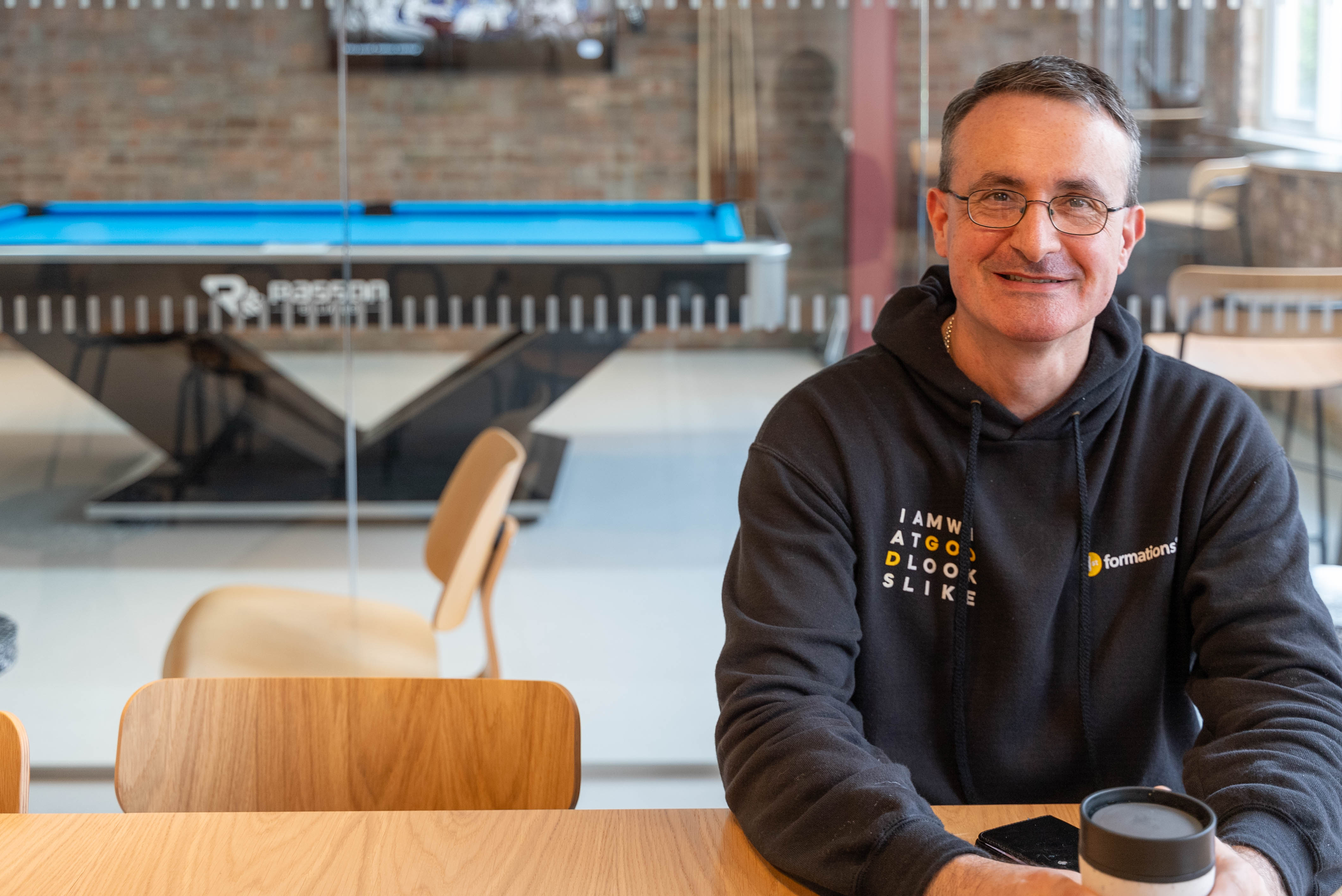 Nick, our Building Manager, sitting at our community table wearing his 1st Formations hoodie and having a coffee, with Games Room in the background.